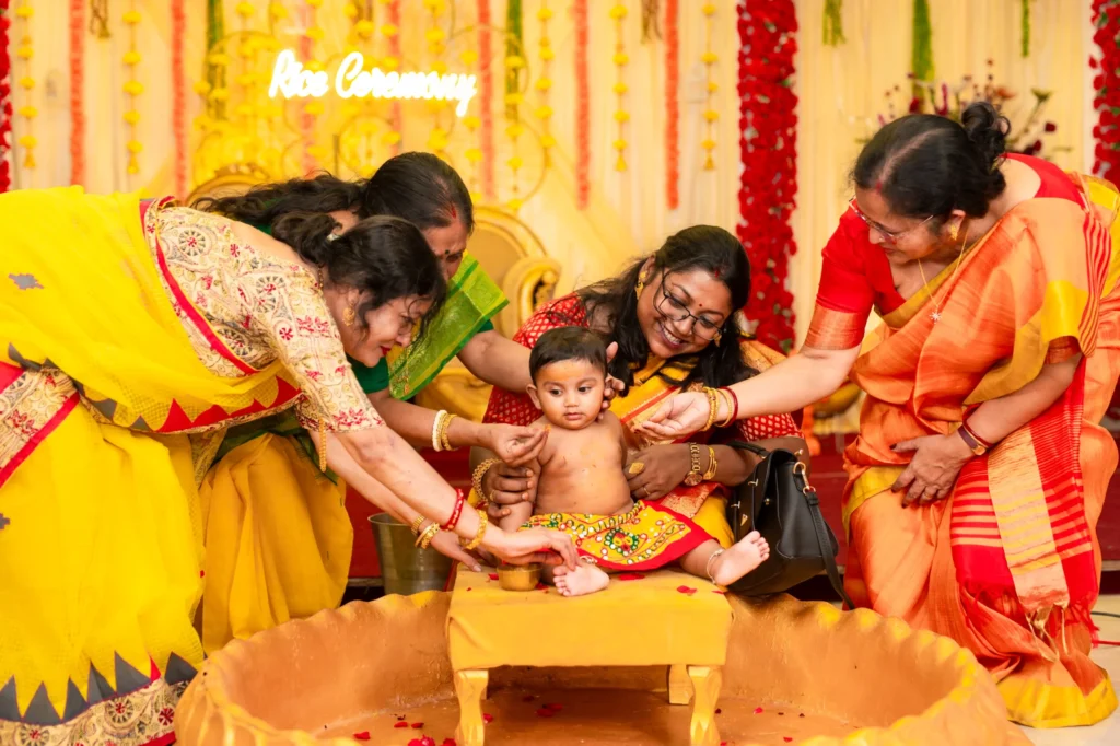 A baby sitting in a traditional Bengali rice ceremony setup, surrounded by family members applying rituals, beautifully captured as part of Bengali rice ceremony photography.